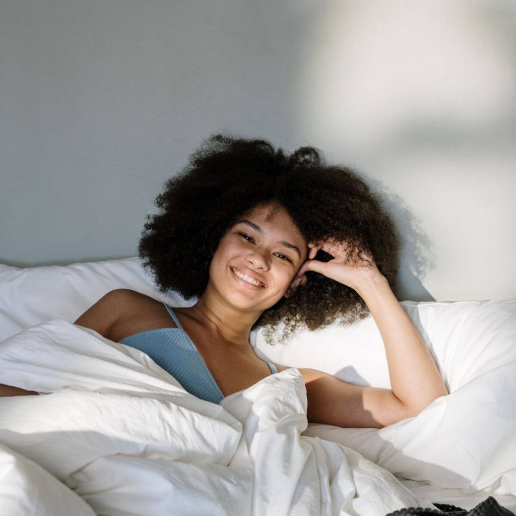An African American woman is laying in bed under a white comforter. She is smiling and wearing a light blue spaghetti strap pajama top.