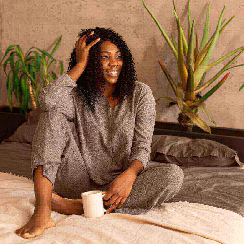 An African American woman is sitting on a bed with grey sheets. She is wearing a dark grey pajamas set and is smiling with a white mug in her hand.