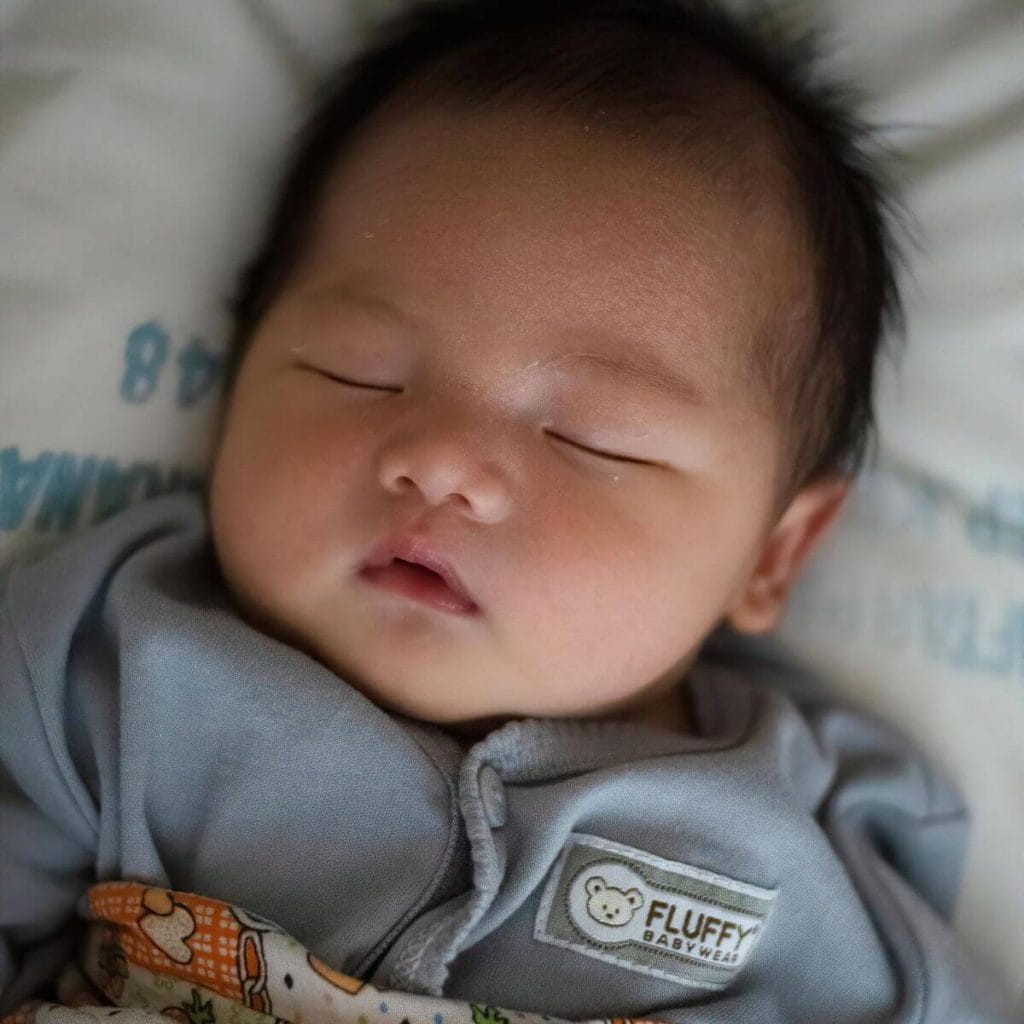 An Asian American newborn boy is laying on a white sheet. He has his eyes closed and is wearing a blue/grey sleep sack