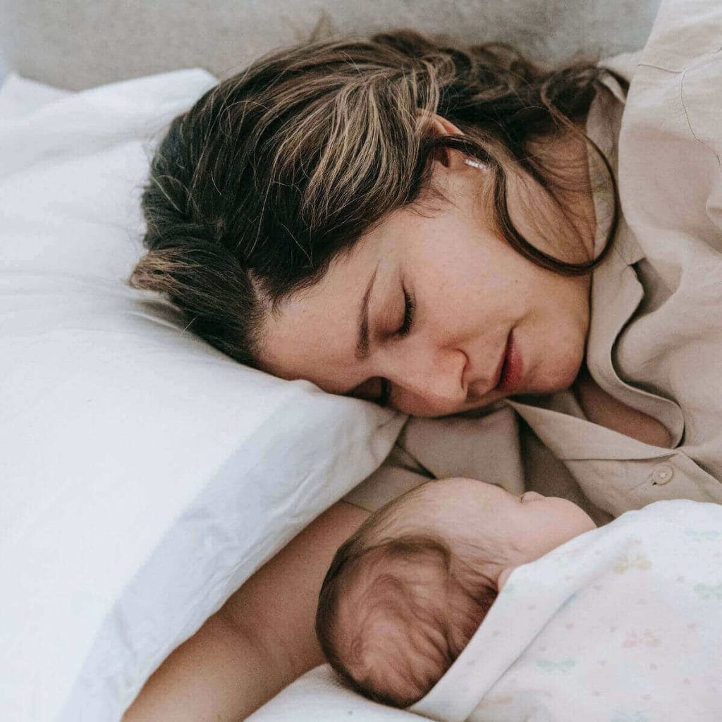 A mom is laying down with her eyes closed with her head on a pillow. A newborn is laying beside her wrapped up and sleeping.