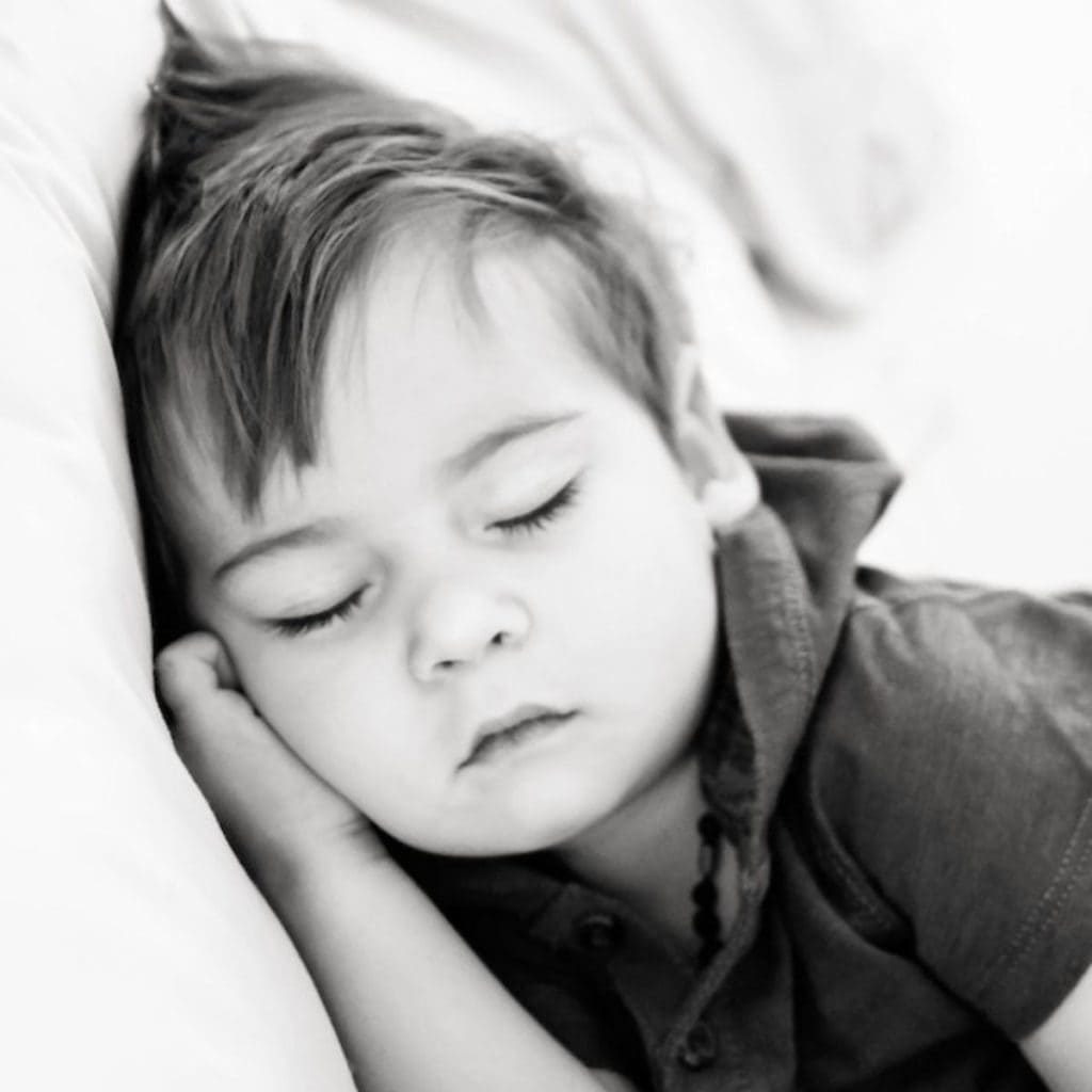 A toddler boy is laying on a white pillow with a collared shirt on. He has his eyes closed and his right hand under his cheek.
