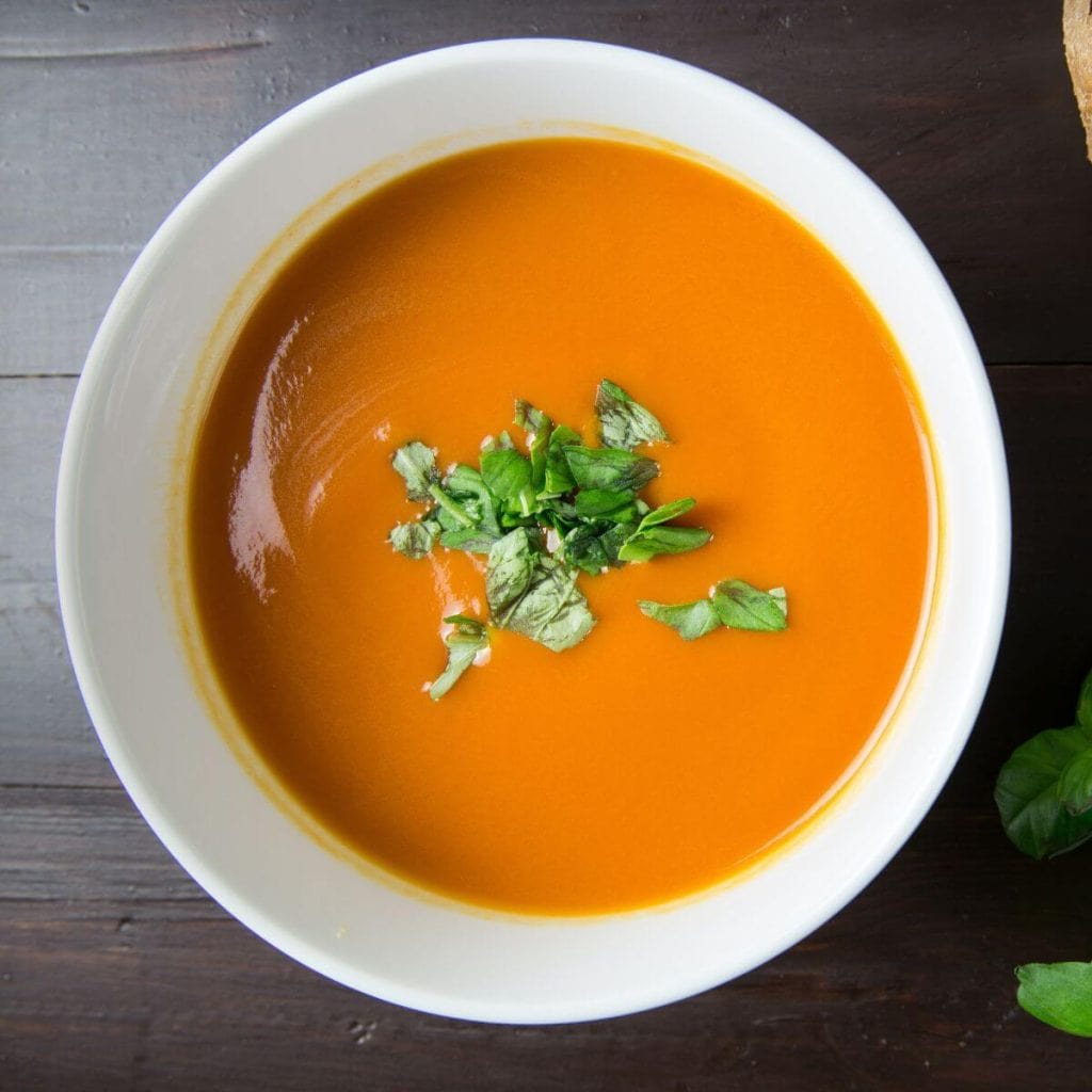On a dark brown wooden table sits a white bowl. Inside the white bowl is a creamy orange soup with green herbs on the top.