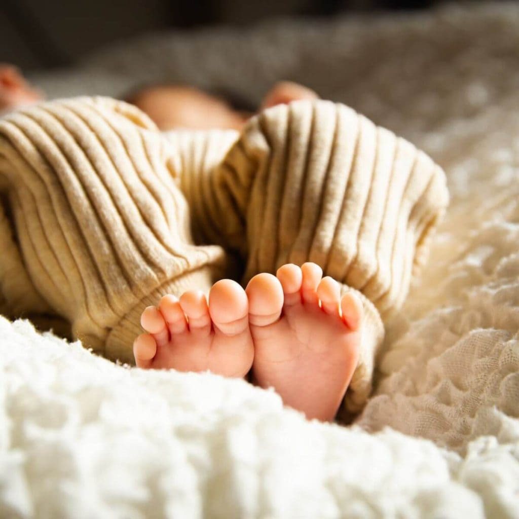 A baby lays on a fluffy white banket in tan colored pajamas with their feet in the center of the picture