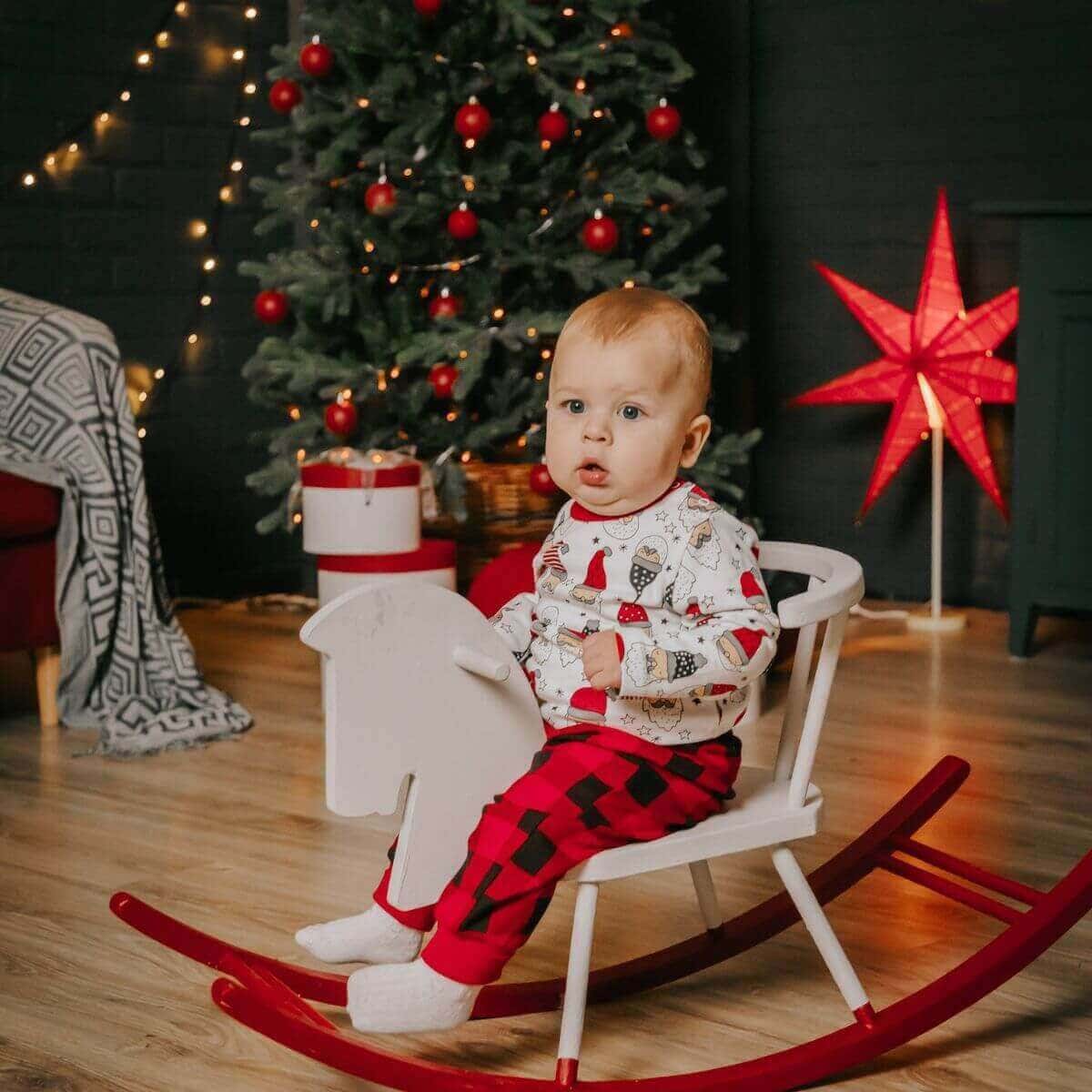 A baby is sitting in a white and red rocking chair in front of a lit up Christmas tree.