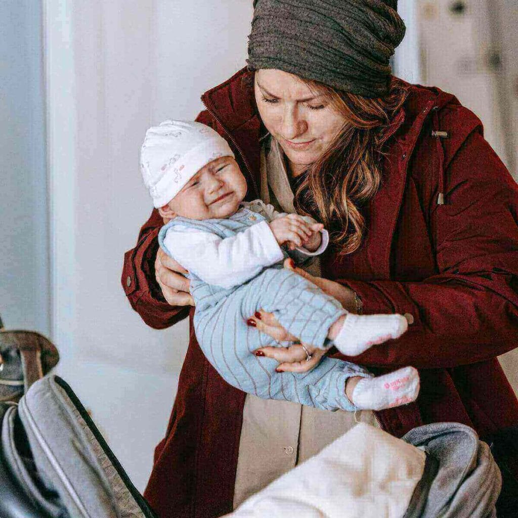 Inside a house, a mom who is dressed in a winter coat and hat is putting her baby dressed in poajamas and a hat into a car seat