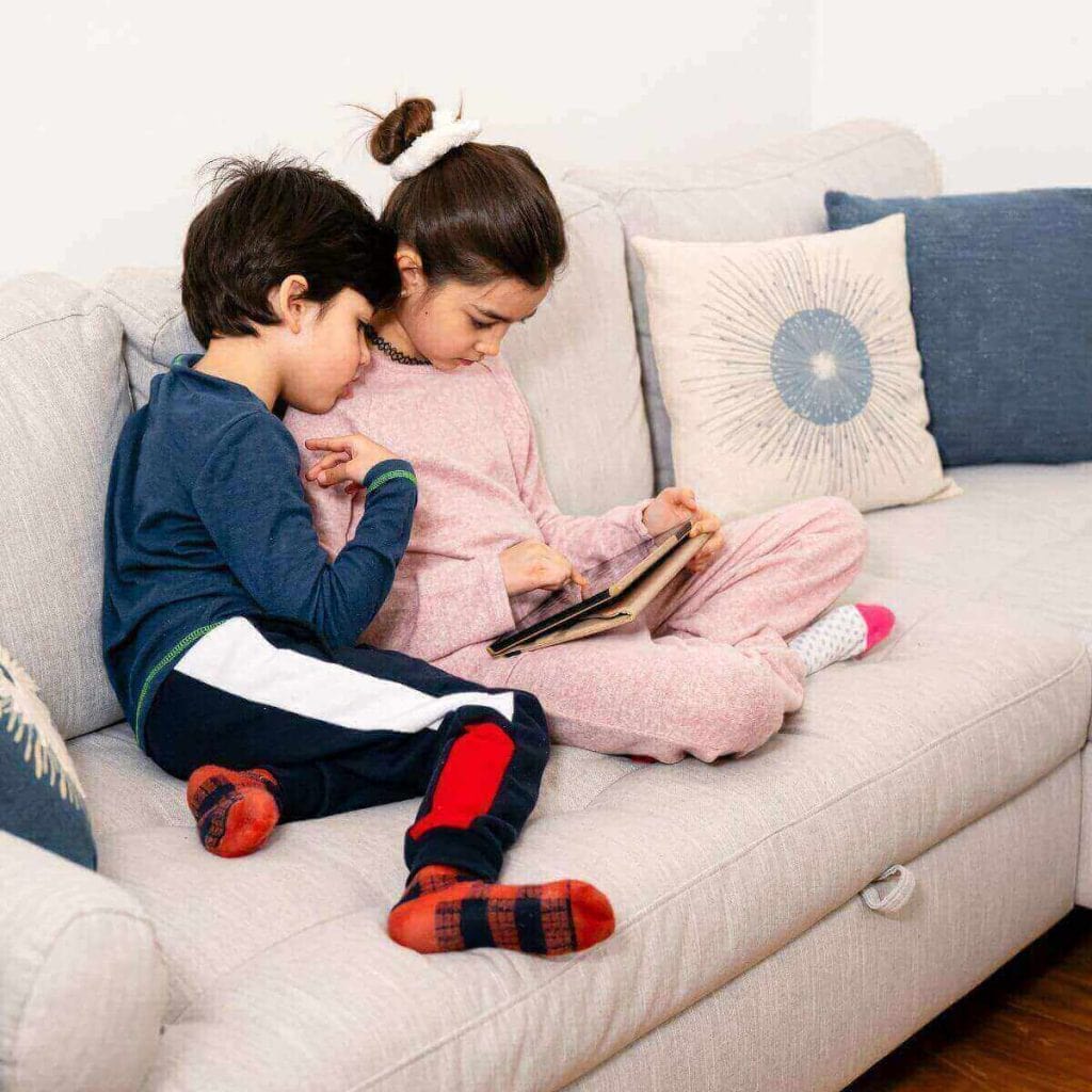 A girl in light pink pajamas and a boy with black,white, and red pants and a dark blue shirt are sitting on a cream colored sofa watching a tablet.
