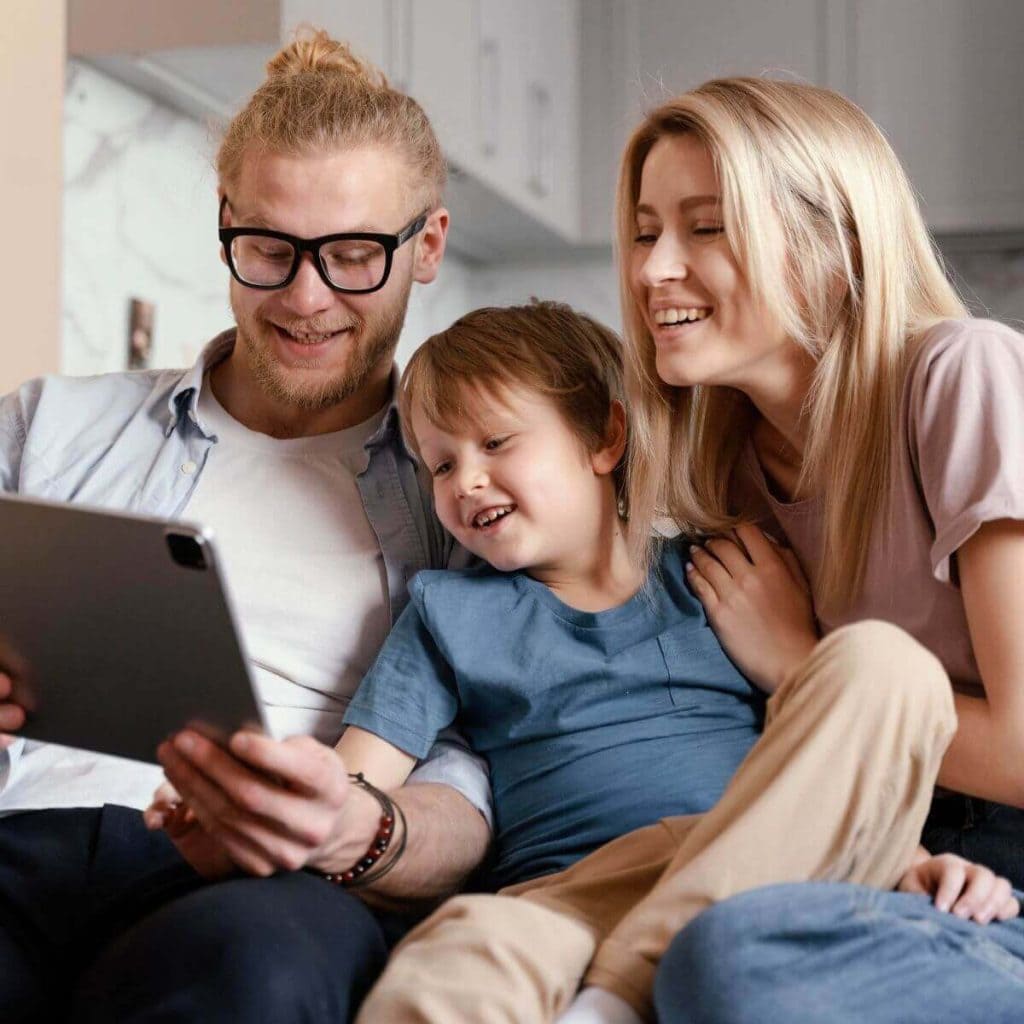 A mom, dad and son are sitting on a couch. The dad is holding a tablet and all three are laughing.