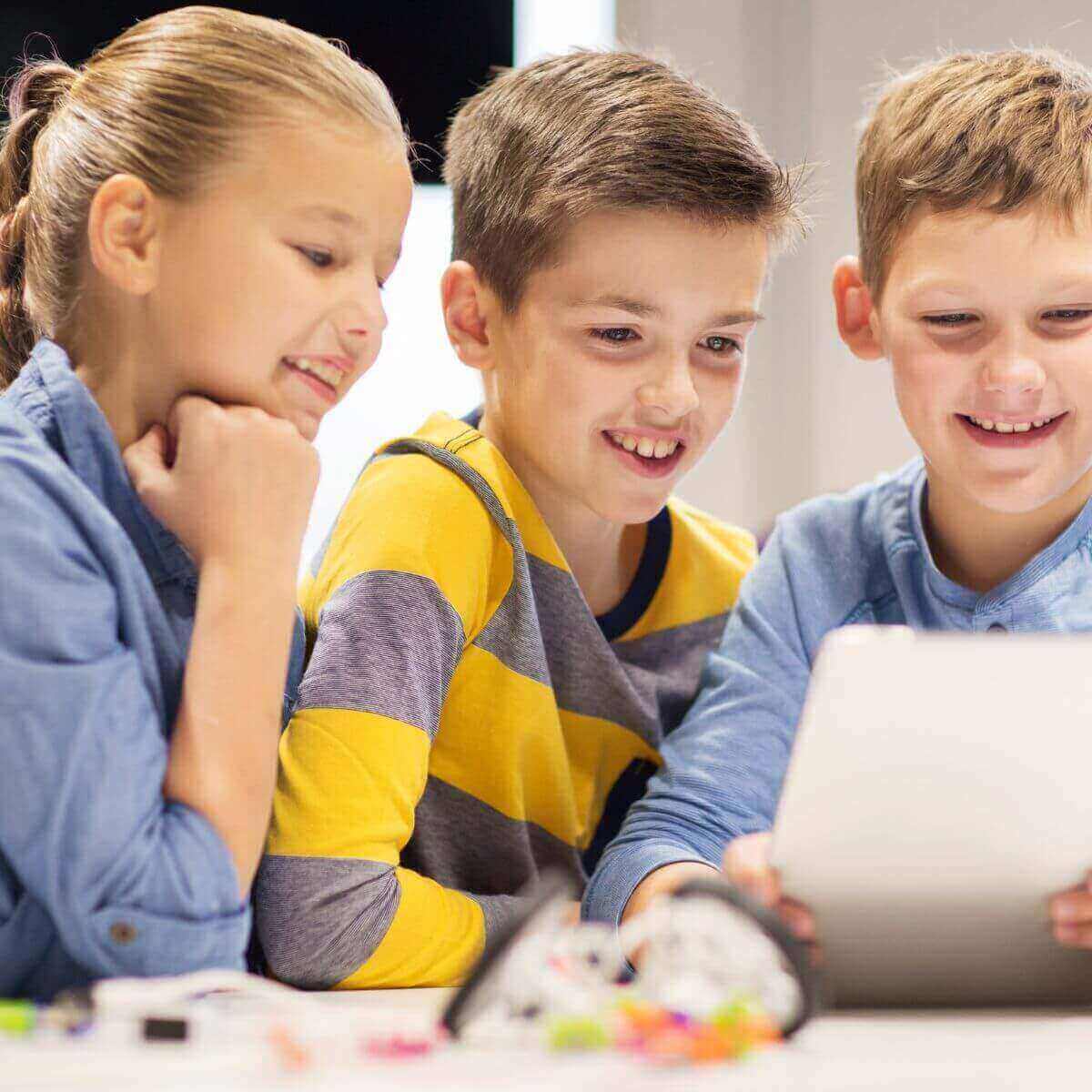 Two boys and a girl are standing at a table watching a tablet and smiling.