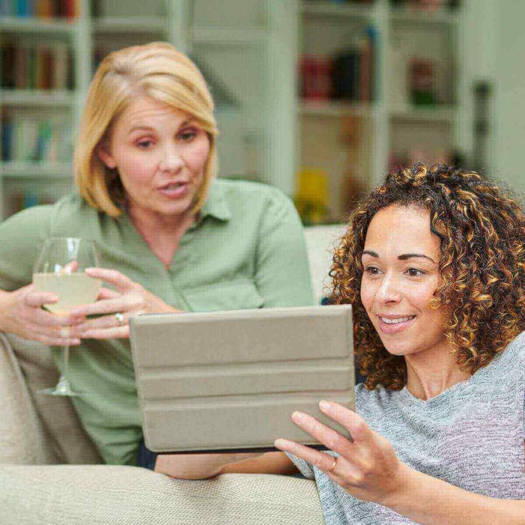 A woman in an olive green shirt is sitting on a couch while a woman in a grey and white shirt is sitting on the floor. They are both looking at a tablet and talking.
