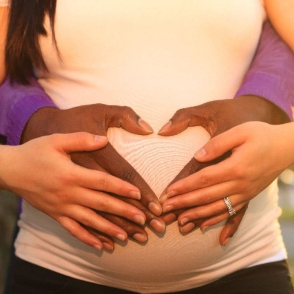 A man and woman are standing up. His hands are in the shape of a heart over her pregnant belly. Her hands are laying on top of his hands.