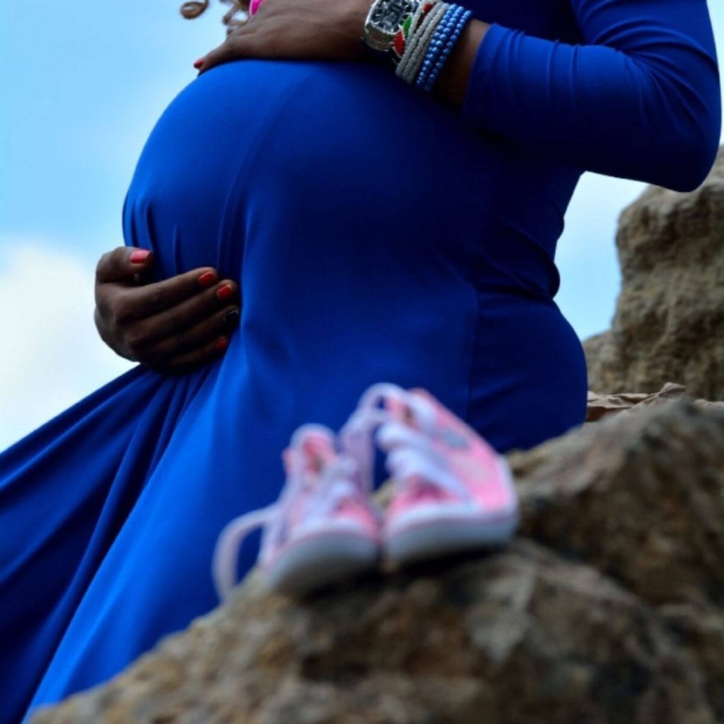 An African American woman is wearing a dark blue dress. She is sitting on big rocks with her left hand on the top of her belly and her right hand below it. There are a pair of pink baby tennis shoes sitting on another rock in front of her.