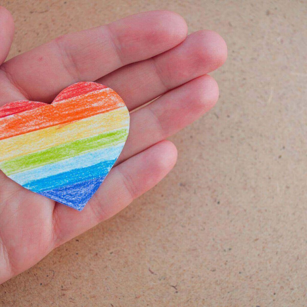 A hand is holding a paper heart with red, orange, yellow, green, light blue, medium blue, and dark blue stripes