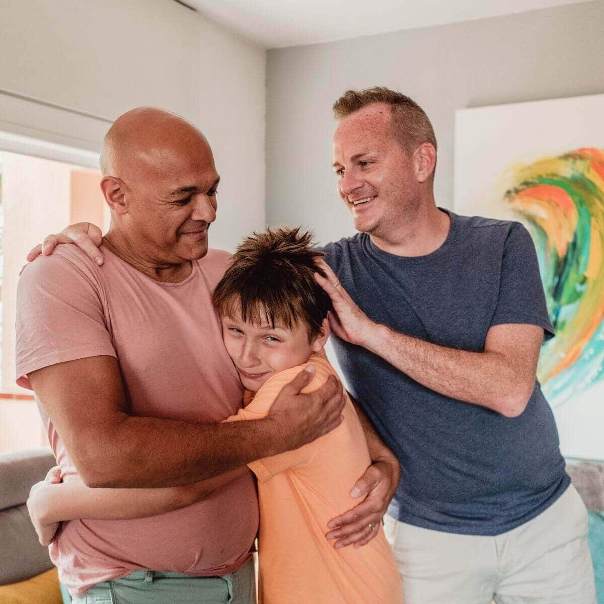 Two men are standing in a living room. One man has his arm around the other man's shoulder while he is hugging a boy that is between them.