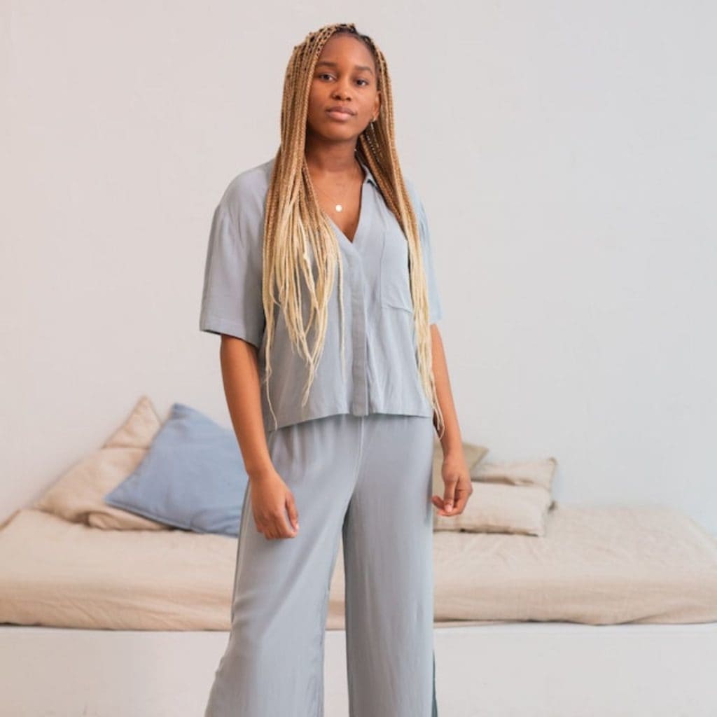 An African American woman is standing in front of a bed. She is wearing light blue bamboo pajamas.