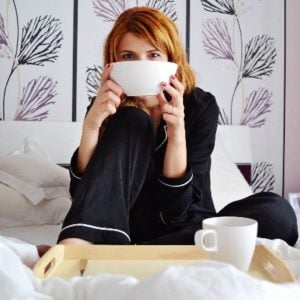 A woman with red hair is sitting on a bed. She is wearing black pajamas and has a white bowl in front of her face.
