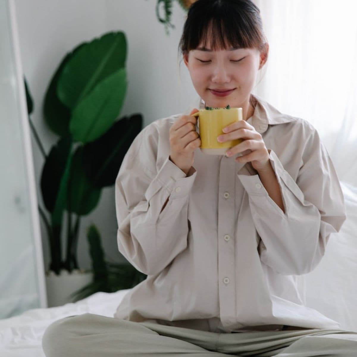 An Asian American woman is sitting on a bed wearing taupe colored pajamas. Her eyes are closed and she is holding a yellow mug in her hands.