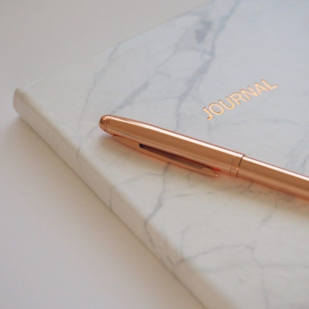 On a white table sits a white marble notebook. On the cover it says "Journal" in rose gold and there is a rose gold pen sitting on top of it.