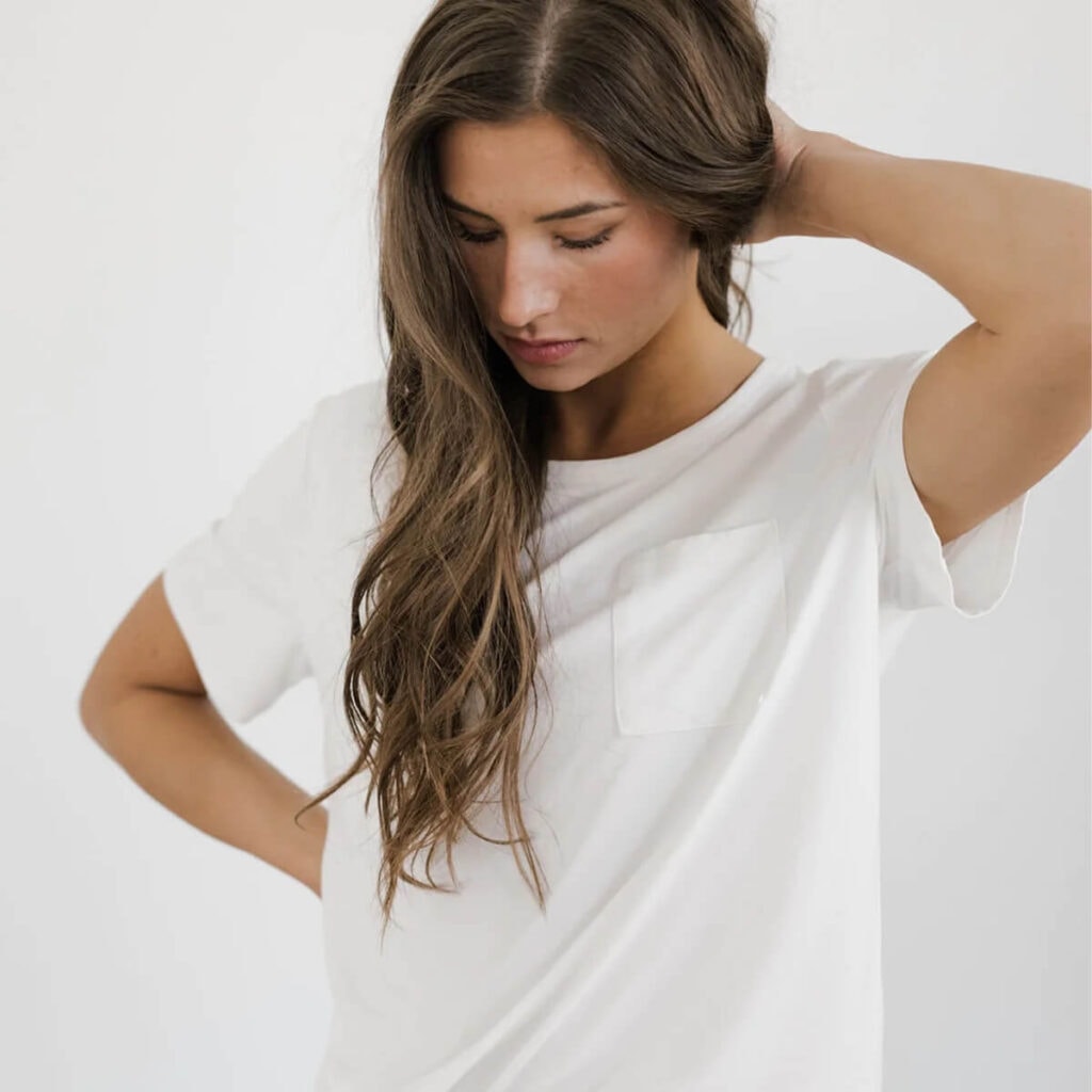 a woman with long brown hair stands in front of a white background wearing a white bamboo shirt. She looks down casually, her face angled away from the camera, with one arm behind her head and the other resting gently on her hip. 