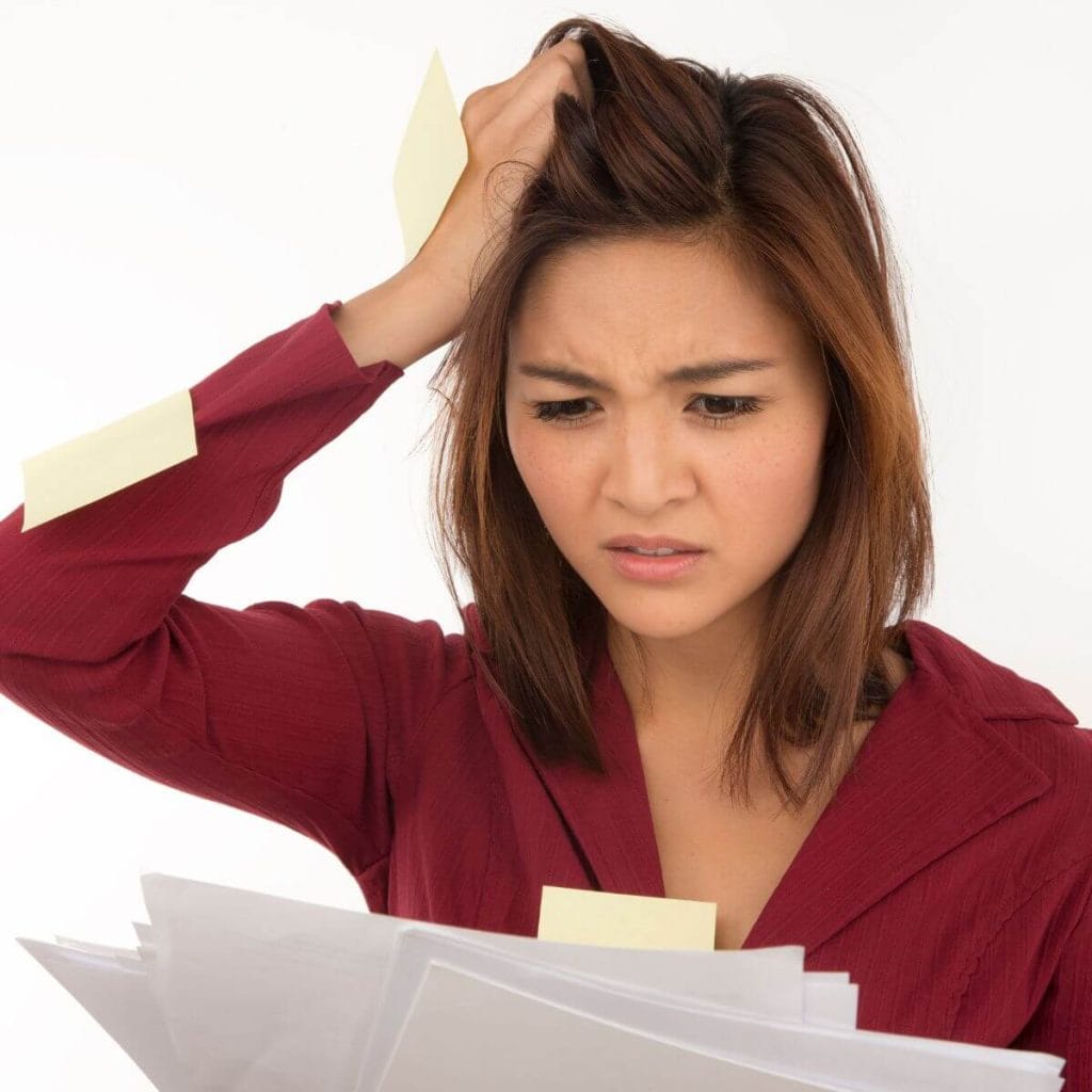 A woman with shoulder length hair is wearing a red long sleeve shirt. She has her right hand in her hair and she is looking down at papers in her left hand like she is confused or overwhelmed.