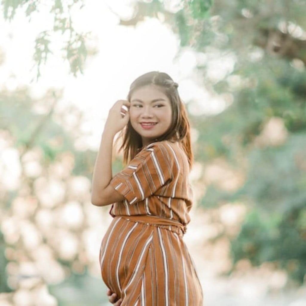 An Asian American woman is standing in a park with trees around her. She is wearing a gold and white striped maternity dress and is smiling.
