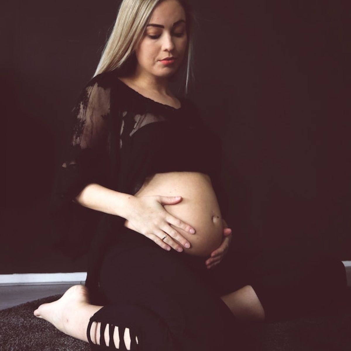A woman is sitting on her knees on a carpeted floor. She is wearing black leggings and a black crop top. She has both hands on her pregnant stomach.