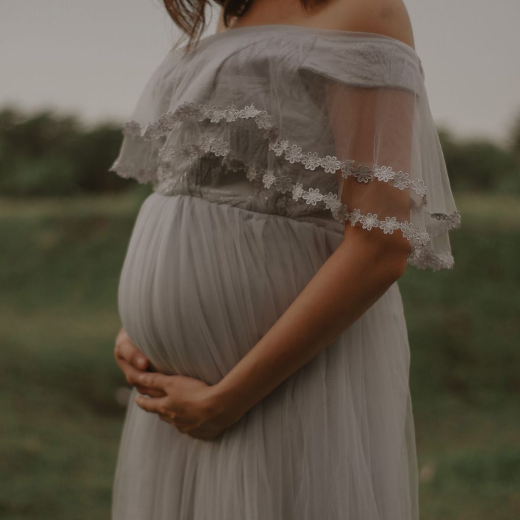 A woman is standing in the middle of a field. She is wearing an off the shoulder white dress and she is holding her pregnant belly with her hands.