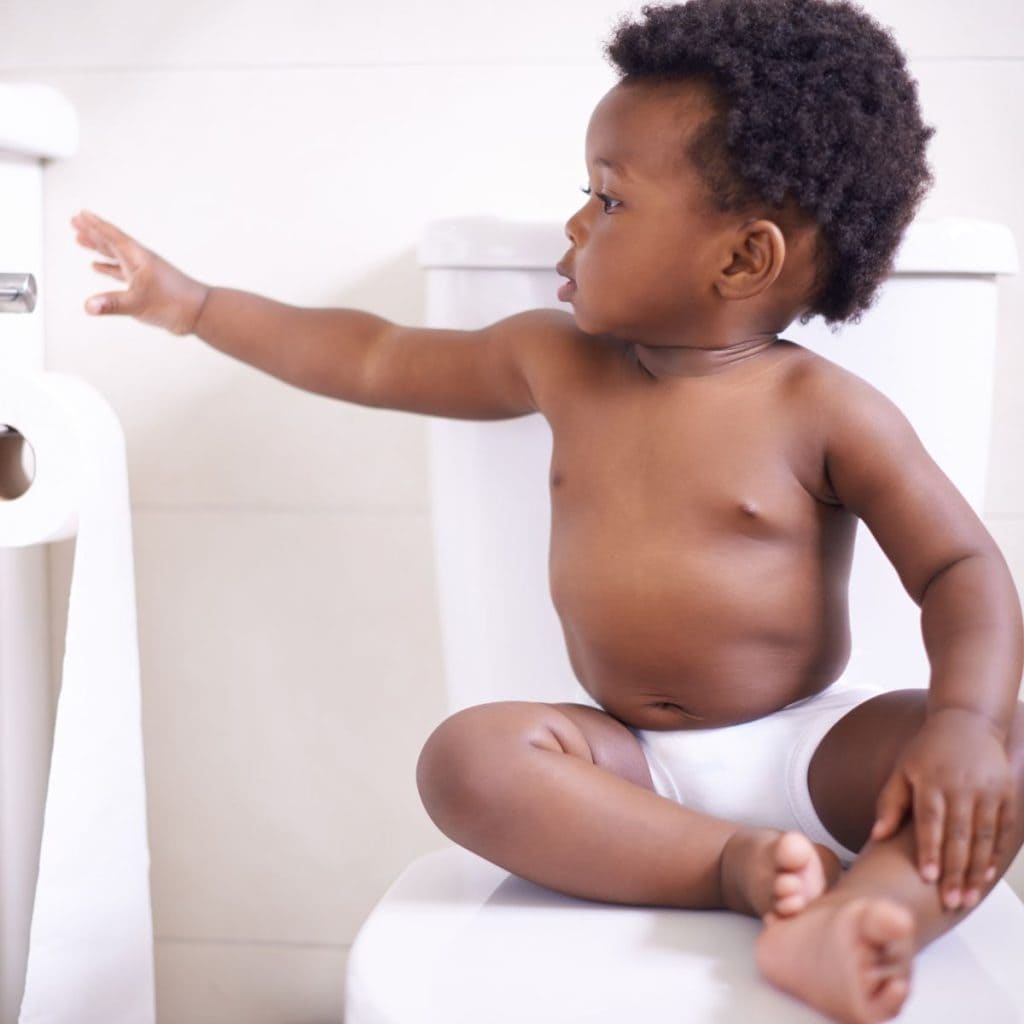 An African American toddler boy is sitting on a toilet in white underwear. He is reaching for a roll of toilet paper