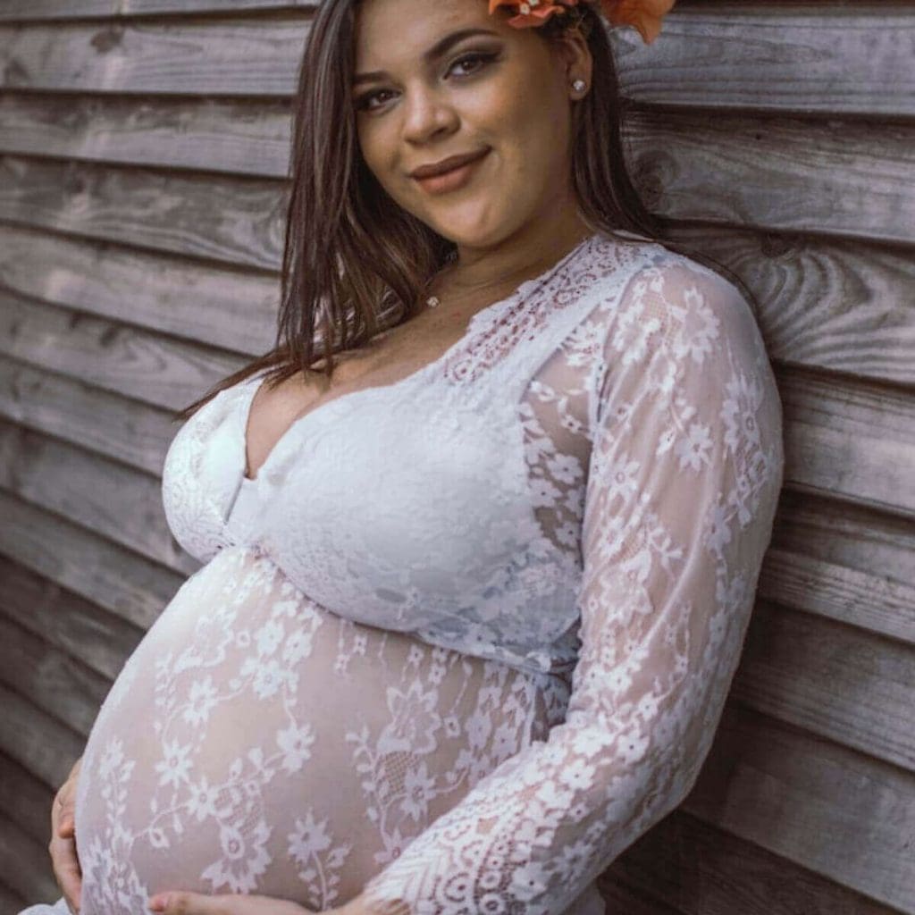 A plus size pregnant woman with long dark brown hair is leaning up against a light grey wood structure. She is wearing a white lace gown and is holding her belly