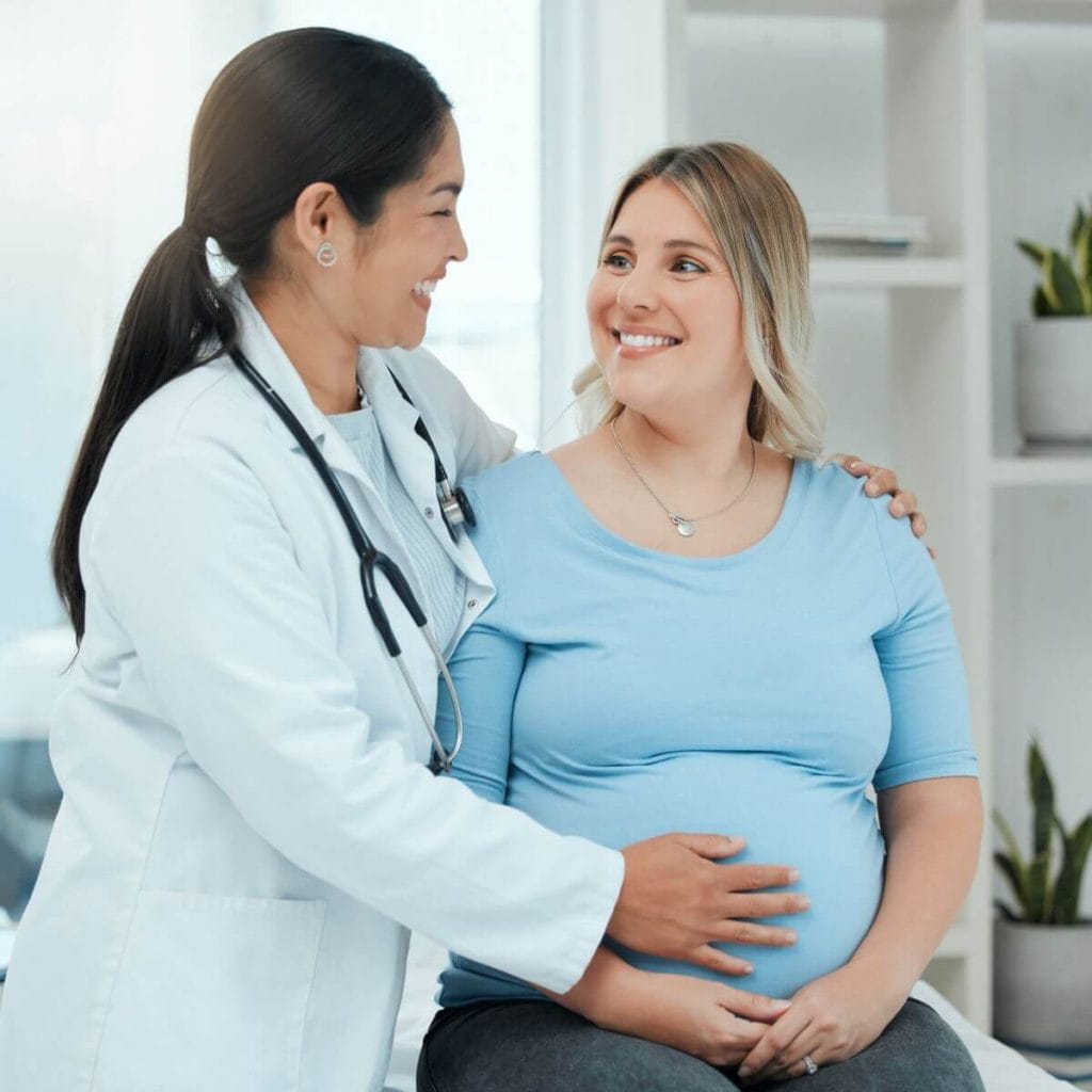 A plus size pregnant woman is sitting on a doctor's table. She is wearing a light blue shirt and the doctor has her hand on her pregnant belly.