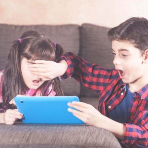 A boy and girl are sitting in front of a couch in a living room. They are looking at a tablet. The boy has his hands over the girls eyes and his mouth has dropped open in disbelief.