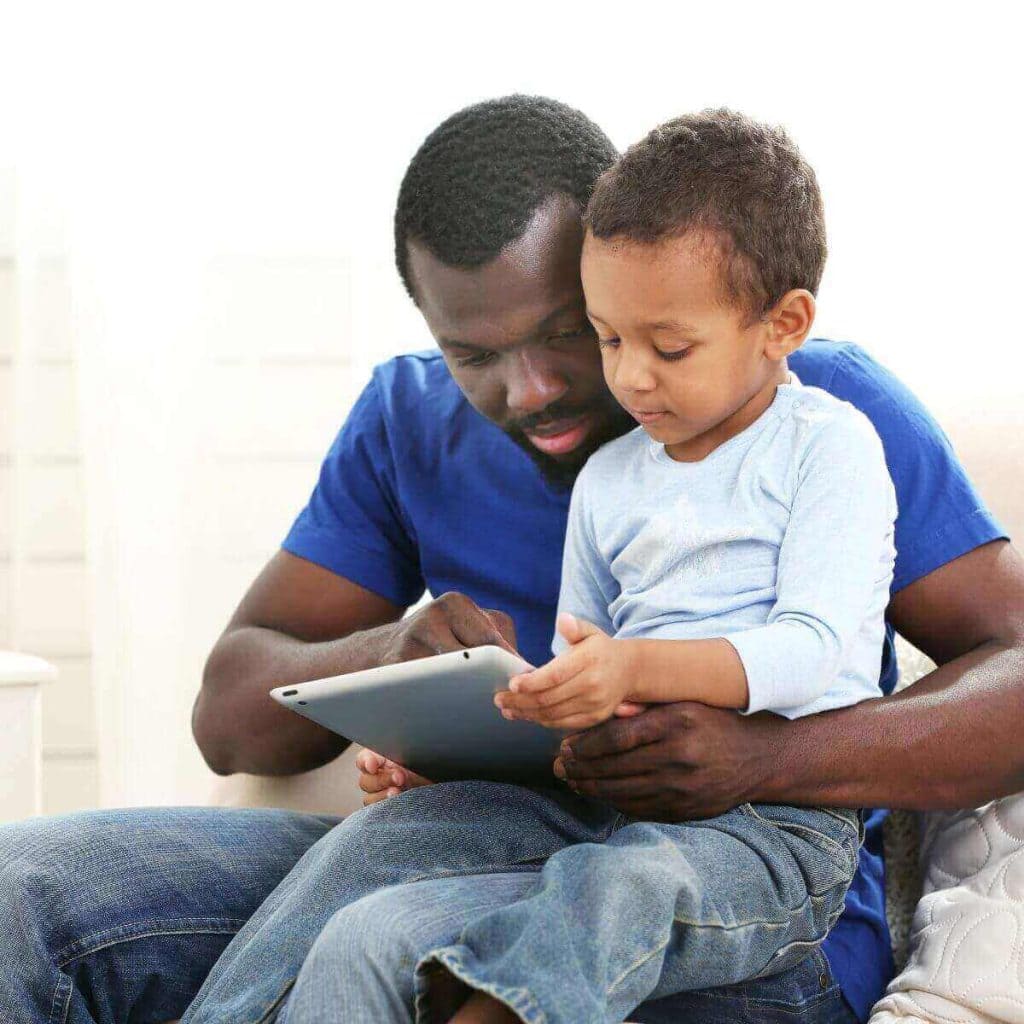 A man in a dark blue shirt and blue jeans has a young boy on his lap. They are looking at a tablet together.