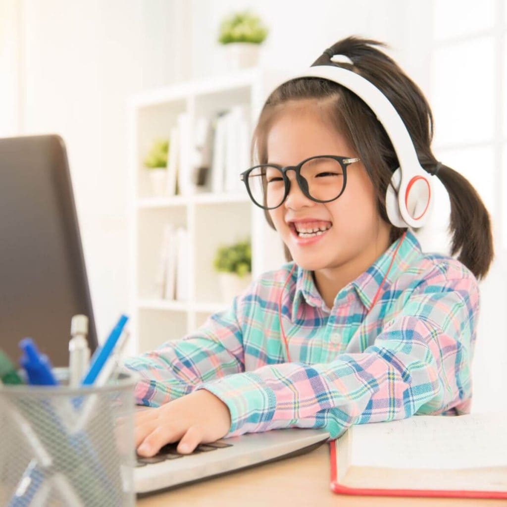 A girl is sitting at a desk in front of a laptop computer. She is wearing black glasses and headphones and she is smiling.