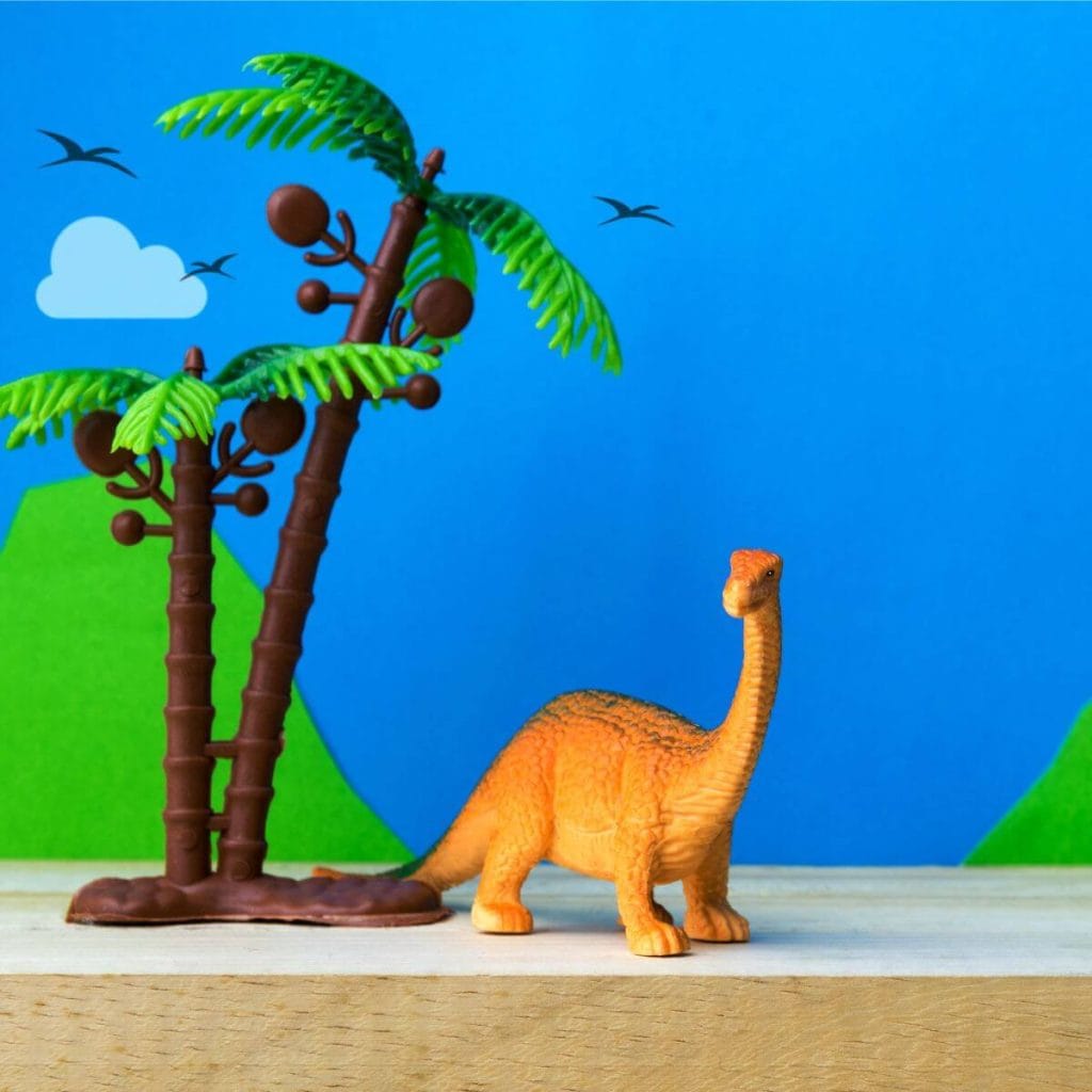 On a white table is an orange long necked dinosaur with two palm trees beside it.
