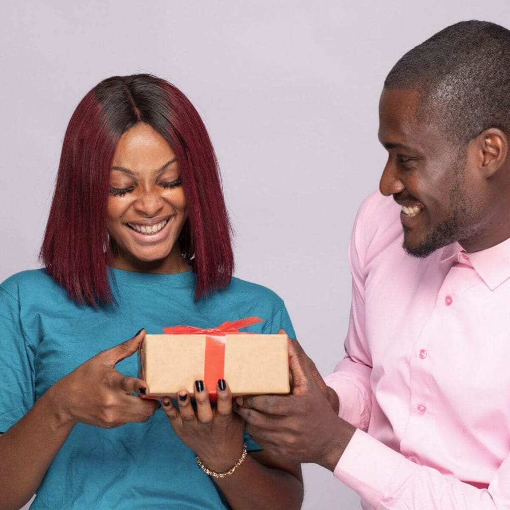 An African American man wearing a light pink shirt is handing a small box to an African American woman wearing a blue shirt.