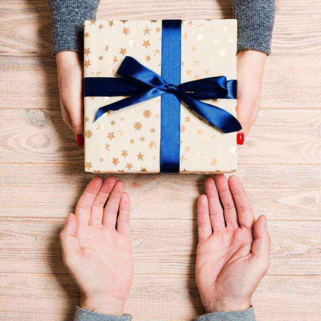 There is a set of man's hands and a set of woman's hands lying on a light wood table. The woman has her hands around a box with cream colored wrapping paper with a blue bow.