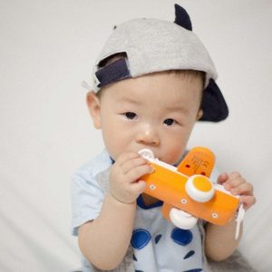 A boy is sitting down. He has a sideways hat on and is holding an orange and white plastic airplane.