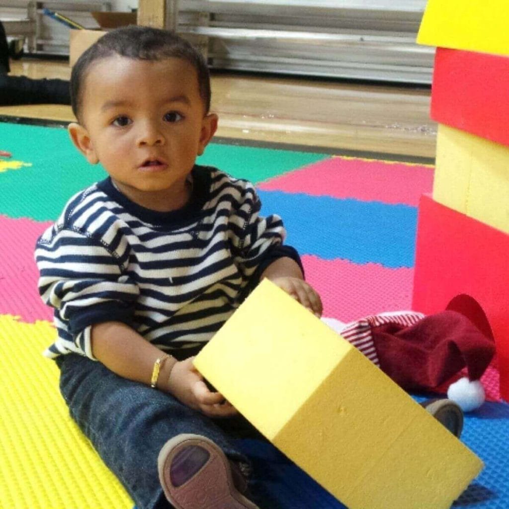 A boy is sitting on red, blue, yellow, and green colored mats. He is in jeans and a blue and white striped shirt and is holding a yellow block.
