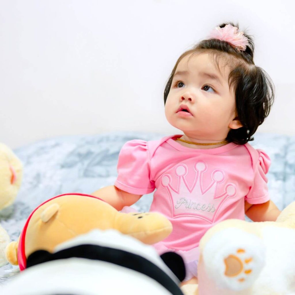 A little girl is sitting on a bed. She is wearing a light pink teeshirt that says princess and she has stuffed animals sitting around her.