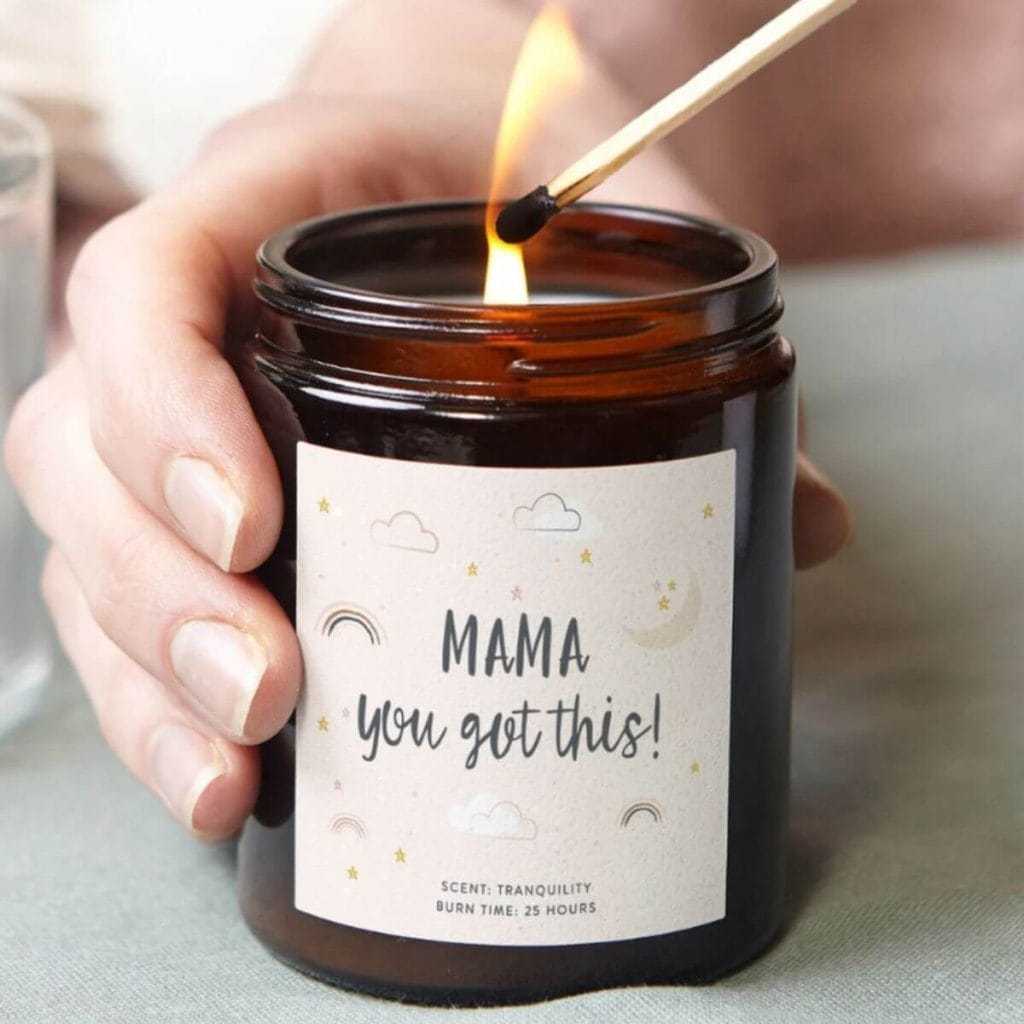A dark brown jar is sitting on a light grey table cloth. Inside the jar is a candle and on the outside it says "Mama You've Got This"