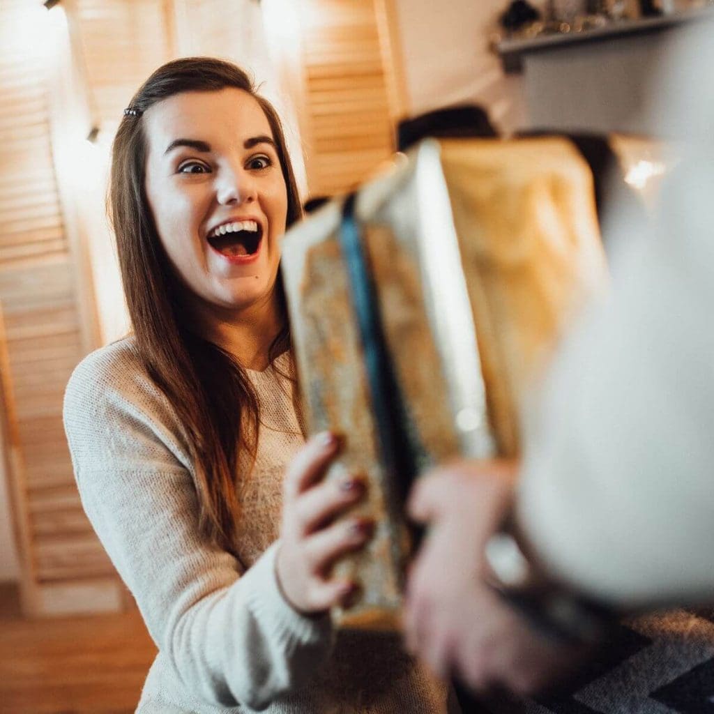 A woman wearing a light brown sweater is sitting on a wooden floor smiling and holding out her hands to get a box wrapped in gold wrapping paper from a man's hands.