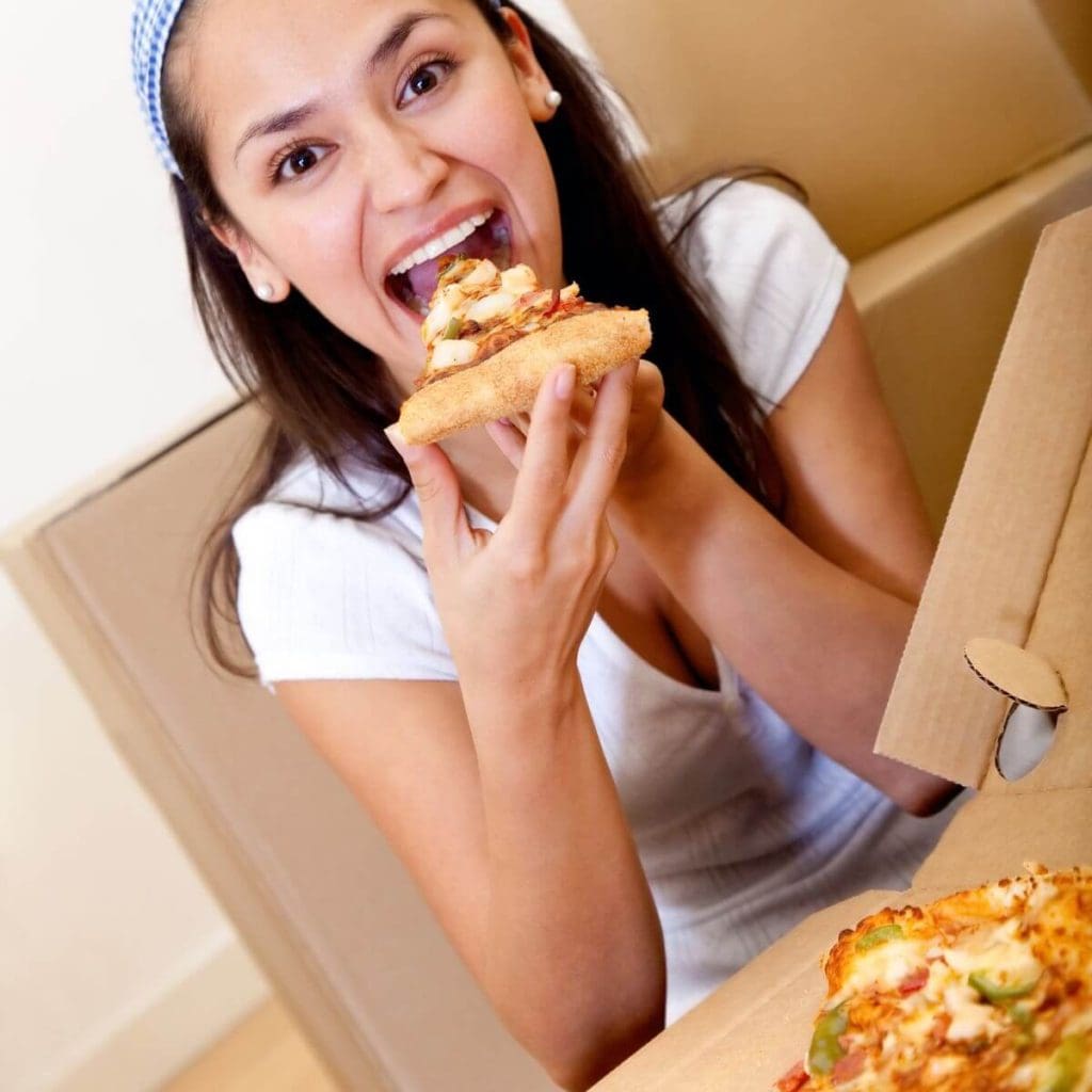 A woman is sitting at a table with a pizza box full of pizza in front of her. She is bringing one slice of pizza up to her mouth to eat.