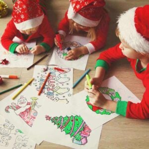 Three kids are laying on their stomach on a hardwood floor. They are wearing red and green Christmas pajamas and santa hats. They are coloring wreaths, Christmas trees, stockings and a Gingerbread man.