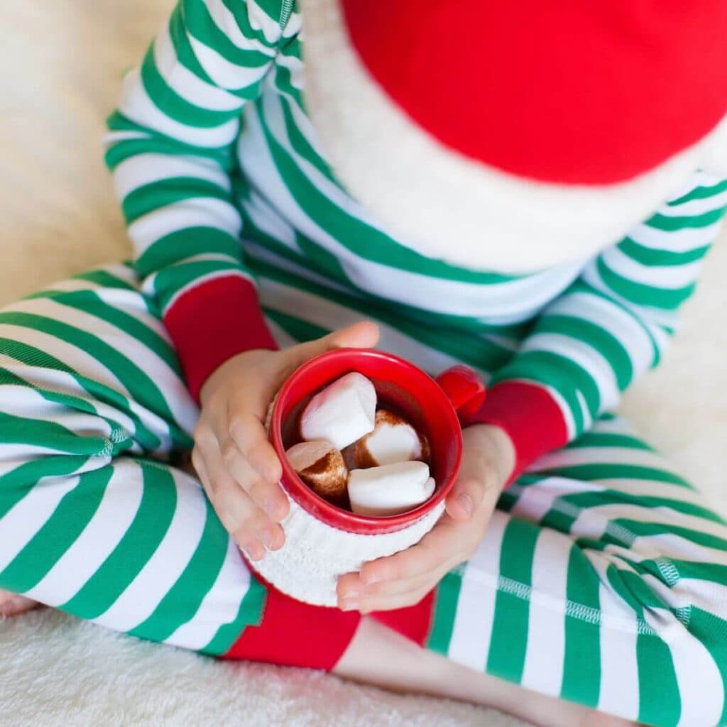 A child in green and white striped pajamas wearing a santa hat is holding a red mug of hot cholocate with 4 huge marshmallows in the top.