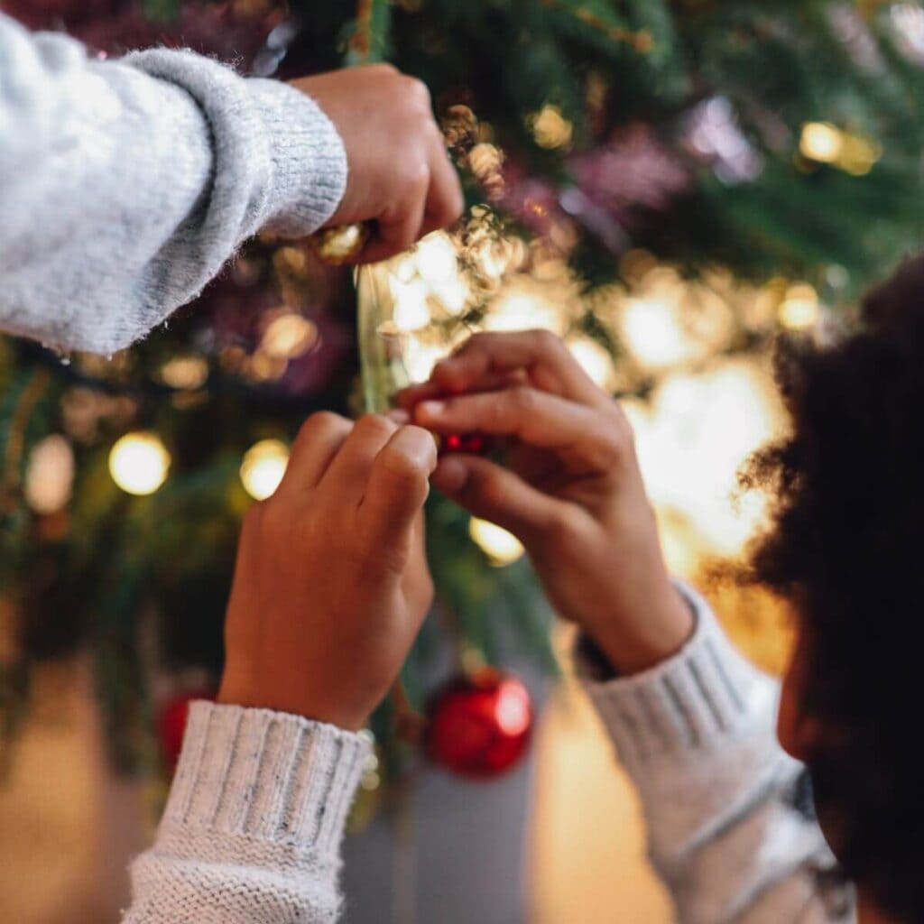 Two children are dressed in light grey sweaters. They are decorating a Christmas tree with red ball ornaments.