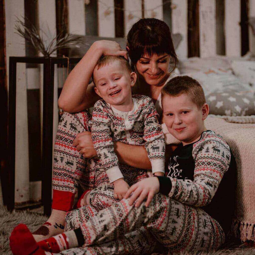 A woman is sitting on the floor beside a bed. Her oldest son is sitting beside her and her younger son is sitting in her lap. All three are wearing the same pajamas that have moose, Christmas trees, and snowflakes.