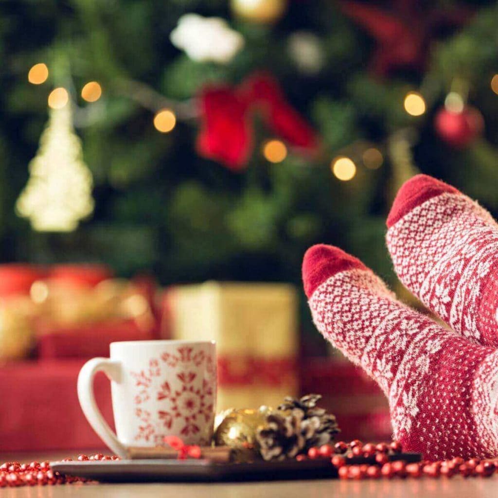A Christmas tree and presents are in the background of the picture. In the front is a white mug with a red snowflake on it and red and white fuzzy socks.