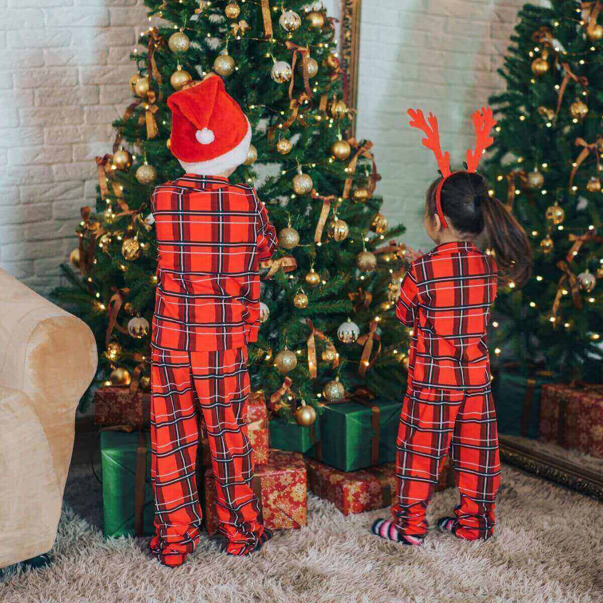 A boy and girl are standing in front of a Christmas tree. Their backs are to the camera. Both are wearing red, black, and white plaid pajamas that match. The boy is wearing a Santa hat and the girl is wearing reindeer ears.