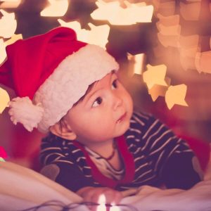 A baby boy is laying on his stomach on a blanket. He is wearing a white and blue stripped shirt and a red santa hat. There are Christmas lights in the background.