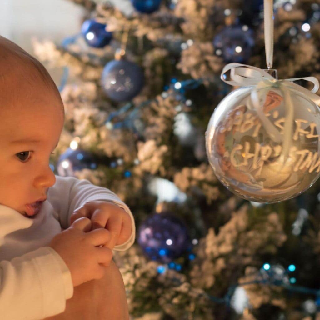 A baby in all white footed pajamas is being held by a man next to a Christmas tree. On the Christmas tree is a silver ball ornament with "Baby's First Christmas" on it.