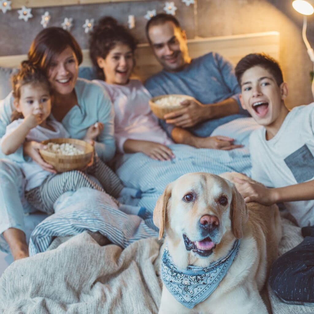 A mom and dad are in a bed with their two daughters, a son, and a dog. There are snowflakes hanging from the wall behind the bed and mom and dad are each holding a bowl of popcorn.